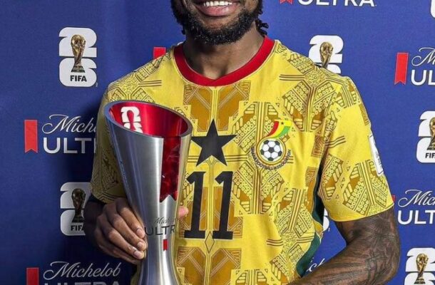 Smiling soccer player in a yellow patterned jersey with number 11, holding a tall silver trophy against a blue Michelob Ultra backdrop.