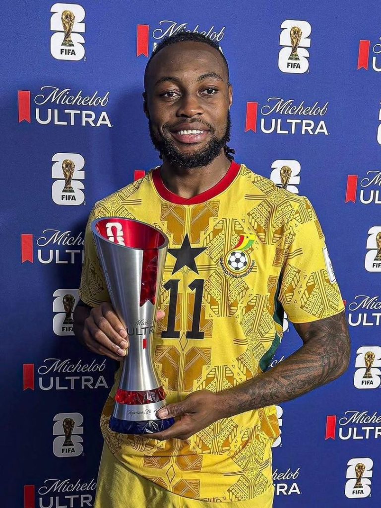 Smiling soccer player in a yellow patterned jersey with number 11, holding a tall silver trophy against a blue Michelob Ultra backdrop.