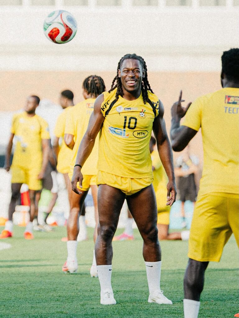 Soccer players in yellow uniforms on a green field; a smiling player with braids watches a colorful ball overhead.