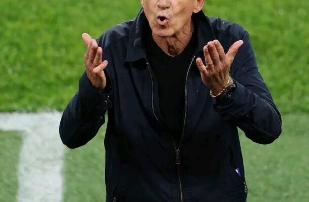 Soccer coach on a green field gesturing emphatically with both hands while speaking to players.