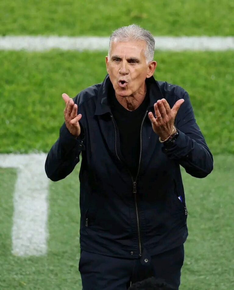 Soccer coach on a green field gesturing emphatically with both hands while speaking to players.