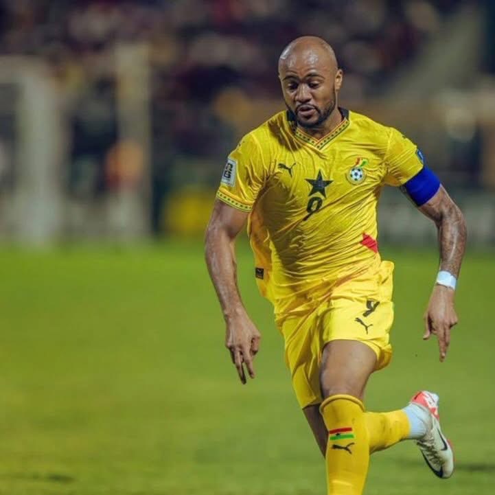Ghana national footballer in yellow kit, blue captain's armband, sprinting on a green pitch during a match.