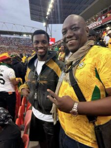 Two smiling fans at a soccer stadium, one in a yellow patterned jersey with scarf and lanyard, the other in a black and yellow jacket.