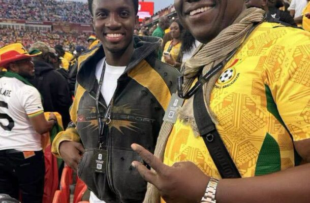 IMG_0995 | Ghanaguardian.com Two smiling fans at a soccer stadium, one in a yellow patterned jersey with scarf and lanyard, the other in a black and yellow jacket.