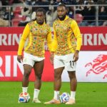 Two soccer players in matching yellow patterned jerseys stand together on a green field with a ball at their feet in a stadium behind red Kirin banners.