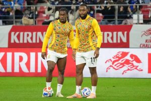 Two soccer players in matching yellow patterned jerseys stand together on a green field with a ball at their feet in a stadium behind red Kirin banners.