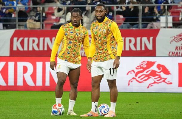 Two soccer players in matching yellow patterned jerseys stand together on a green field with a ball at their feet in a stadium behind red Kirin banners.