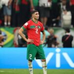 Soccer player in red jersey and green shorts standing on a pitch with hands on hips, crowd in the background (Portugal team).