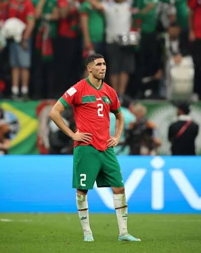 Soccer player in red jersey and green shorts standing on a pitch with hands on hips, crowd in the background (Portugal team).