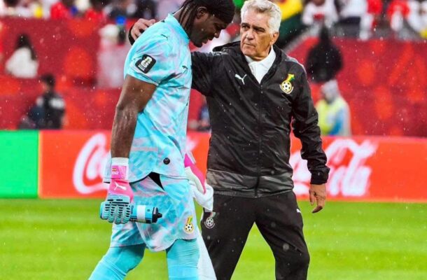 Soccer goalkeeper in light-blue kit walks with a coach or trainer draping an arm over his shoulder on a rainy field, stands filled with fans in the background.