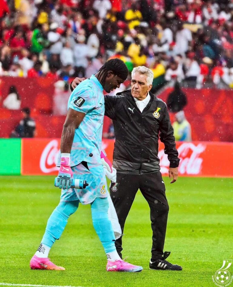Soccer goalkeeper in light-blue kit walks with a coach or trainer draping an arm over his shoulder on a rainy field, stands filled with fans in the background.