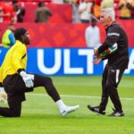 Soccer coach hands a ball to a kneeling goalkeeper in a bright yellow kit during training on a grass field, stands and spectators in the background.