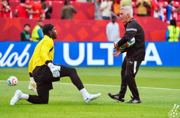 Soccer coach hands a ball to a kneeling goalkeeper in a bright yellow kit during training on a grass field, stands and spectators in the background.