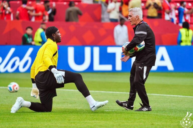 Soccer coach hands a ball to a kneeling goalkeeper in a bright yellow kit during training on a grass field, stands and spectators in the background.
