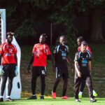 Soccer players stand by the goalpost on a sunny green field, wearing red and black uniforms, ready for a match or practice.