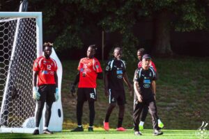 Soccer players stand by the goalpost on a sunny green field, wearing red and black uniforms, ready for a match or practice.