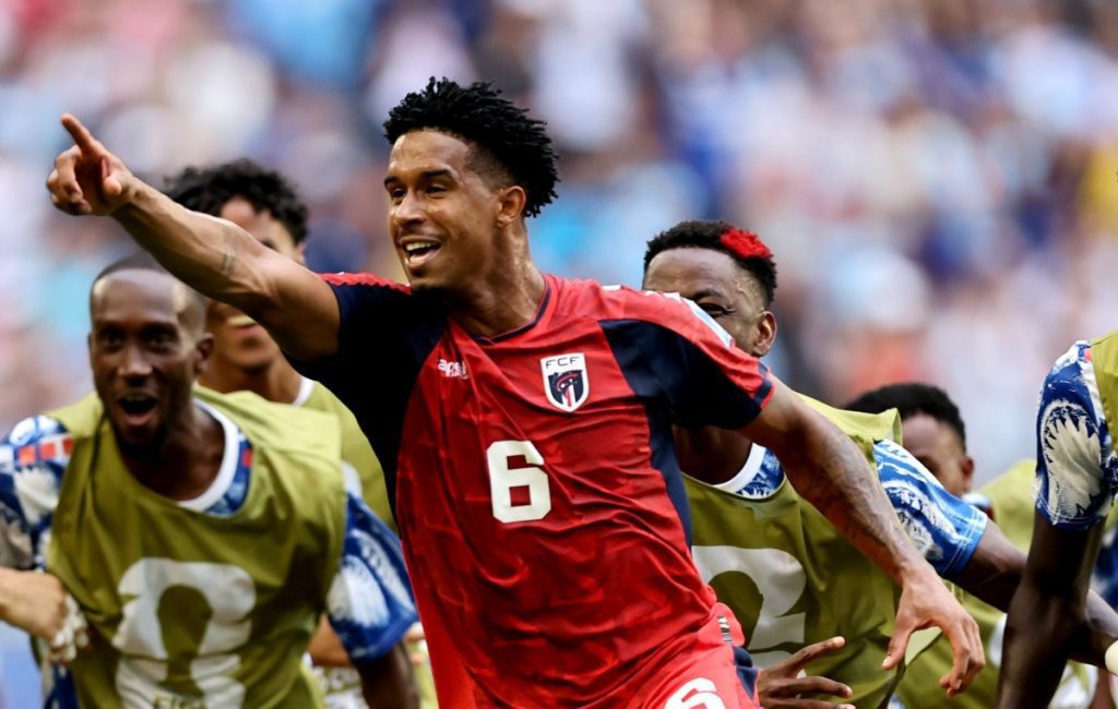 Soccer player in a red jersey number 6 raises his right arm in celebration as teammates in yellow bibs surround him on the field.