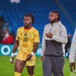 Two Ghanaian soccer players walk on the field after a match; left player in a yellow jersey with number 10, right in a gray hoodie.