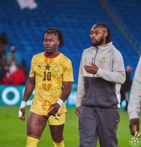 Two Ghanaian soccer players walk on the field after a match; left player in a yellow jersey with number 10, right in a gray hoodie.