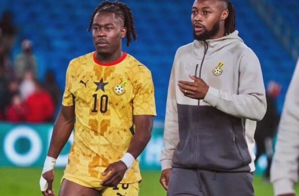 Two Ghanaian soccer players walk on the field after a match; left player in a yellow jersey with number 10, right in a gray hoodie.