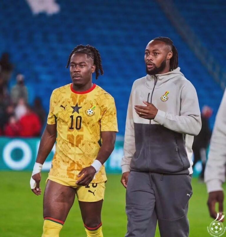 Two Ghanaian soccer players walk on the field after a match; left player in a yellow jersey with number 10, right in a gray hoodie.