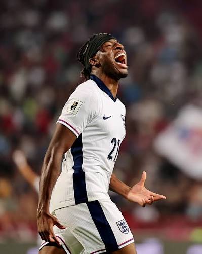 England national team soccer player in a white kit shouting in celebration on the pitch, arms outstretched.