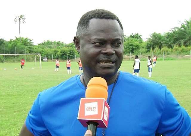 Man in a blue shirt speaks into an orange KWESA microphone on a soccer field, with players practicing in the background.