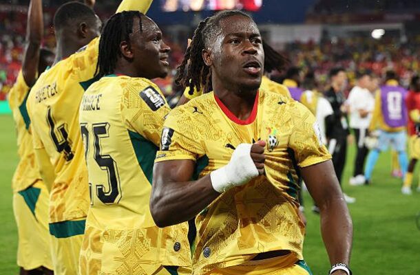 Jamaican national football players celebrate on the field; foreground player clenches his fist, wearing number 15 on his back, others cheer in yellow jerseys.