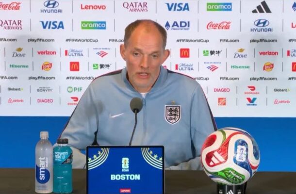 England national team coach speaking at a press conference with a blue backdrop full of sponsor logos, a microphone in front, a branded soccer ball on the table, and water bottles nearby.