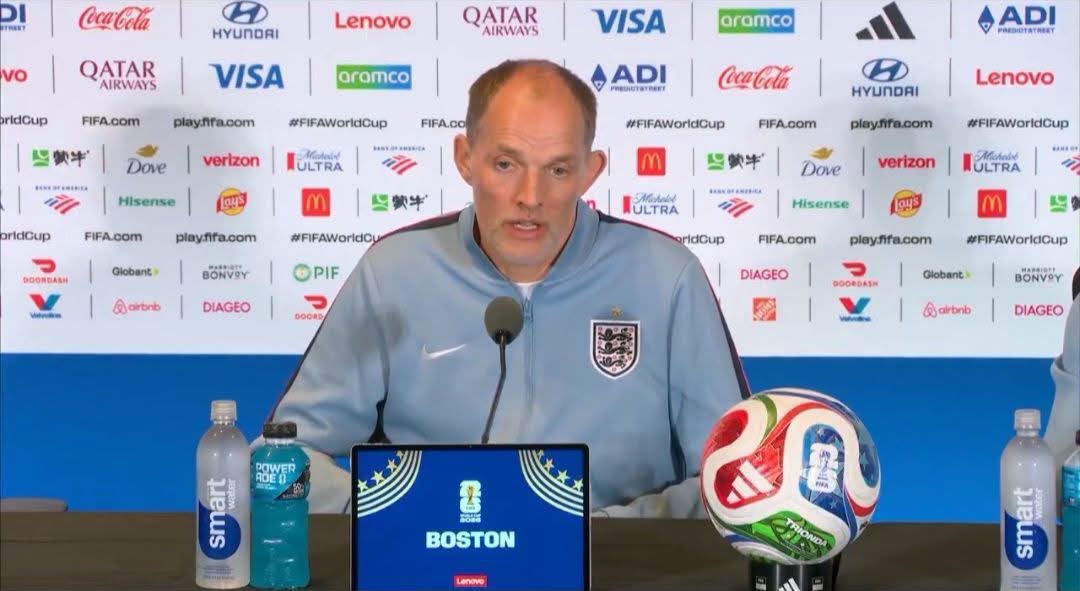 England national team coach speaking at a press conference with a blue backdrop full of sponsor logos, a microphone in front, a branded soccer ball on the table, and water bottles nearby.