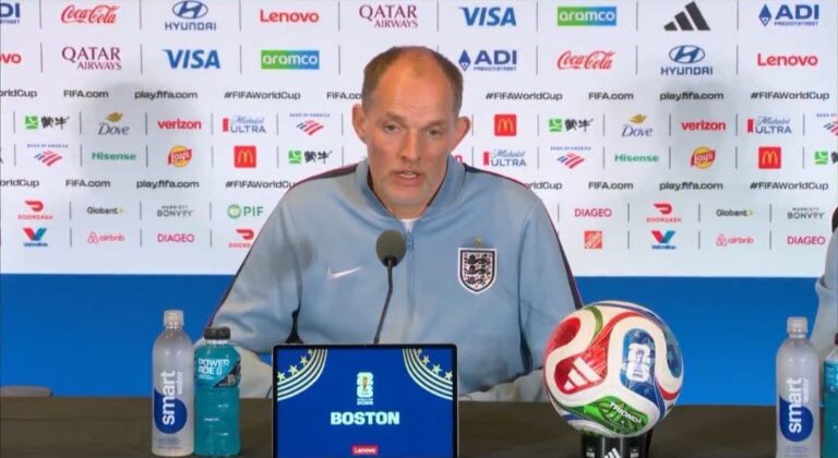 England football manager at a press conference, speaking into a microphone, wearing a light blue jacket with the England crest, with a colorful World Cup ball on the table and sponsor backdrop behind him