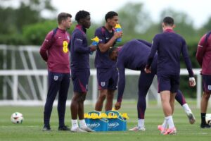 England footballers in maroon training kits stand on a grass field, some drinking water during a training session with equipment nearby.