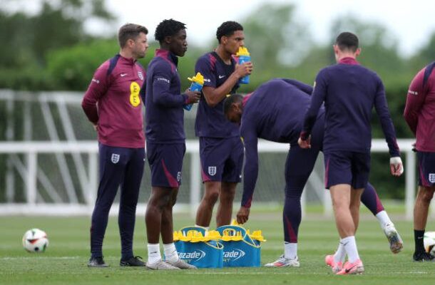 England footballers in maroon training kits stand on a grass field, some drinking water during a training session with equipment nearby.