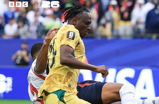 Soccer player in a yellow jersey is challenged by an opponent during a match, with a crowded stadium in the background and BBC/FIFA logos in the corner.