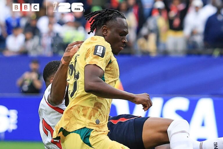 Soccer player in a yellow jersey is challenged by an opponent during a match, with a crowded stadium in the background and BBC/FIFA logos in the corner.