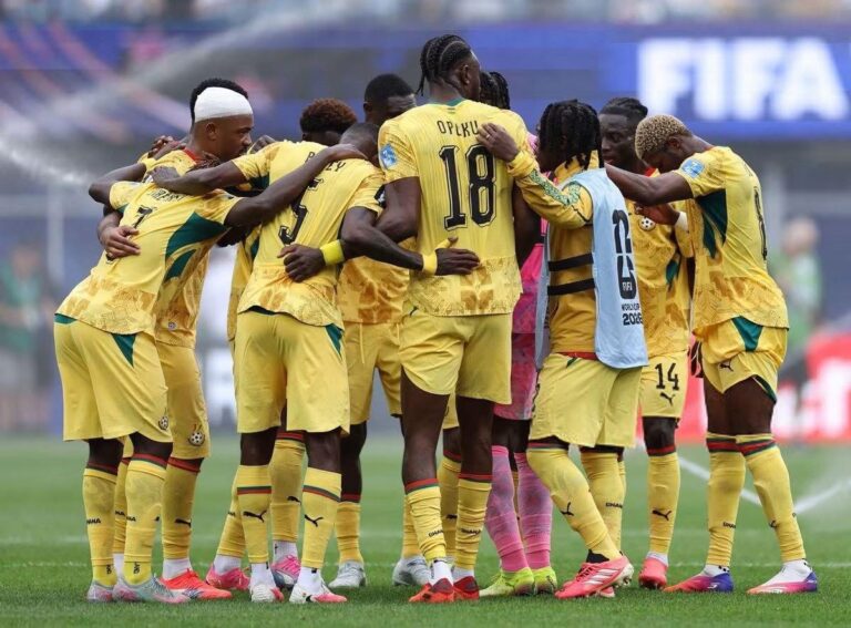 Soccer team in yellow jerseys huddles together on a green field before a match, players with numbered backs forming a circle behind the goalie in pink kit
