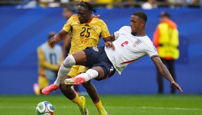 Two soccer players clash for the ball mid-field, one in a white jersey and the other in yellow, both stretching for control as a ball rolls ahead on the grass.