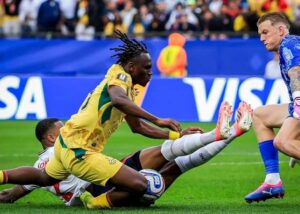 Soccer players in yellow and white contest the ball on a grass field during a match, with one player sliding to intercept the ball.