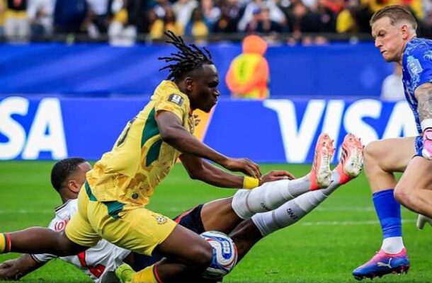 Soccer players in yellow and white contest the ball on a grass field during a match, with one player sliding to intercept the ball.