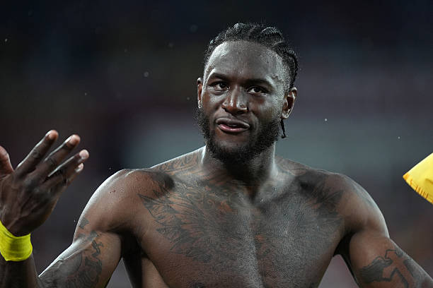 Shirtless athletic man with cornrow braids and chest tattoos gesturing on a sports field in a dim setting.