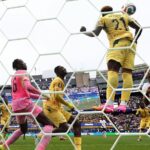 Soccer goalkeeper in pink kit blocks a shot as players in yellow contest near the goal net.