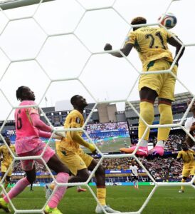 Soccer goalkeeper in pink kit blocks a shot as players in yellow contest near the goal net.