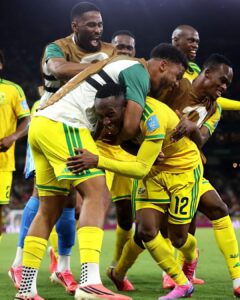 Soccer players in yellow and green celebrate a goal, embracing each other on the field during a match at night.