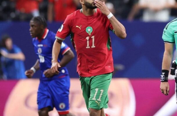 Moroccan footballer in red jersey #11 and green shorts blowing a kiss while running on a soccer field.
