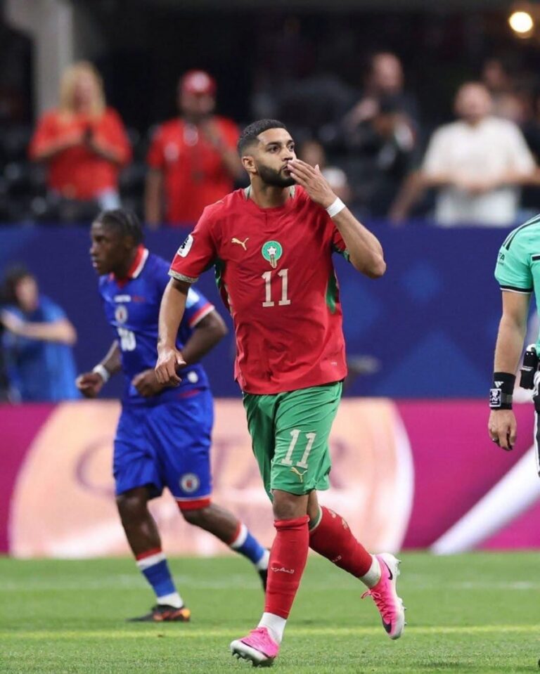 Moroccan footballer in red jersey #11 and green shorts blowing a kiss while running on a soccer field.