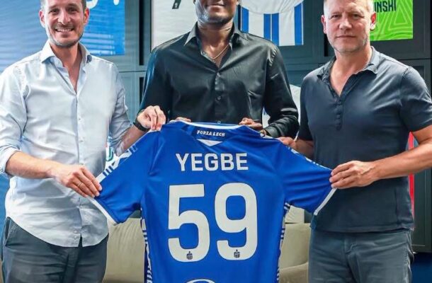 Three men pose in a locker-room setting, presenting a blue soccer jersey with the name 'YEGBE' and number 59 on the back, while framed jerseys hang on the wall in the background.