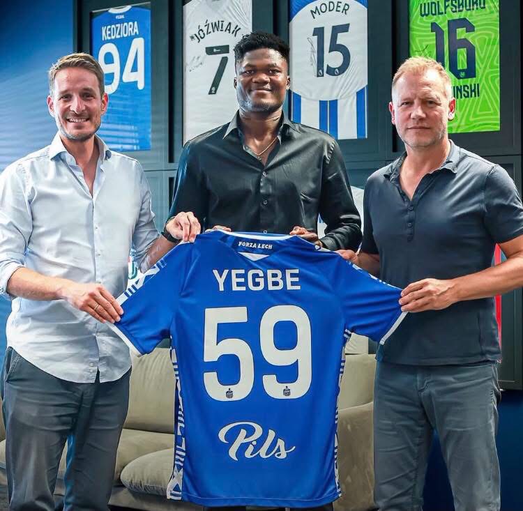 Three men pose in a locker-room setting, presenting a blue soccer jersey with the name 'YEGBE' and number 59 on the back, while framed jerseys hang on the wall in the background.