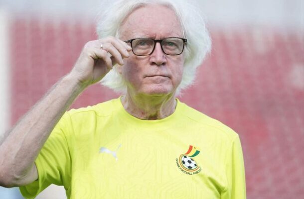 Older man with white hair wearing glasses adjusts his frame while looking at the camera, in a bright yellow football jersey with a Cameroon badge on the chest.