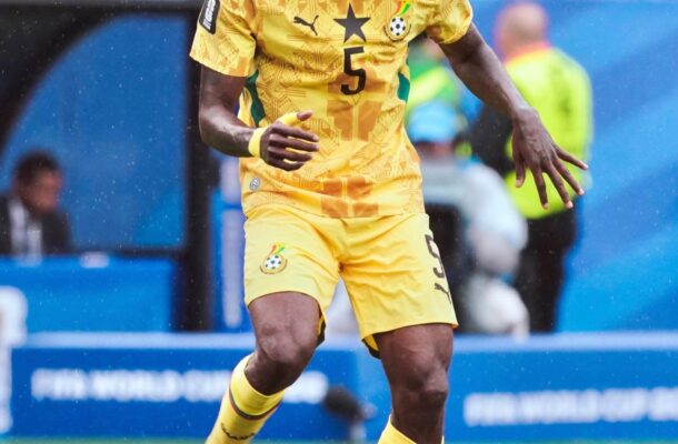 Ghana footballer in yellow kit dribbling a ball on a green field with a crowd watching.