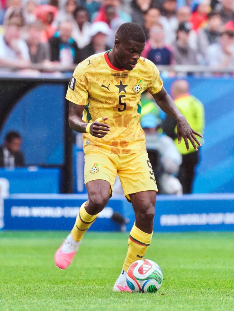 Ghana footballer in yellow kit dribbling a ball on a green field with a crowd watching.